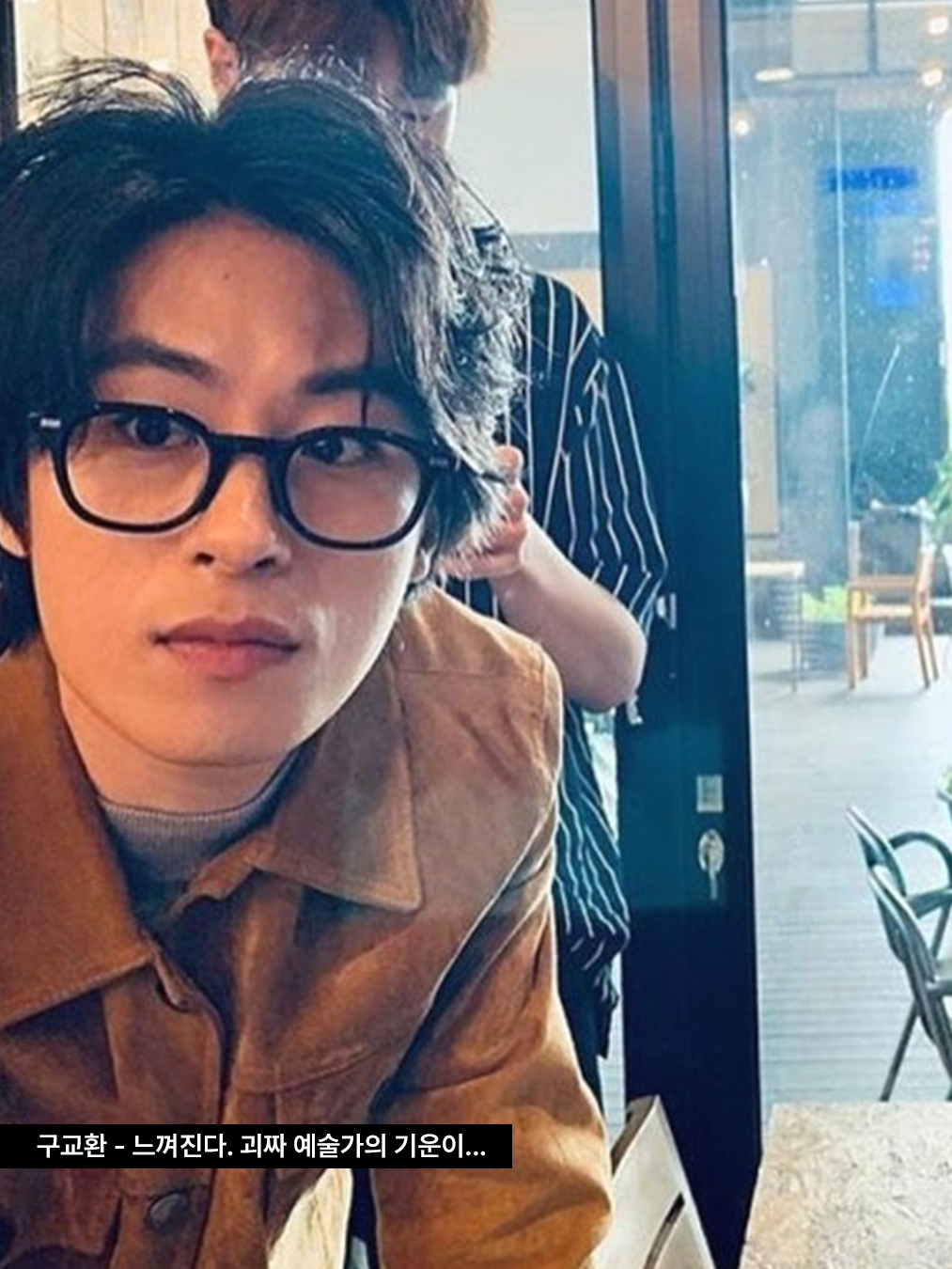 굵은 프레임의 검은 뿔테 안경과 그레이 티셔츠, 브라운 스웨이드 재킷을 입은 배우 구교환의 사진