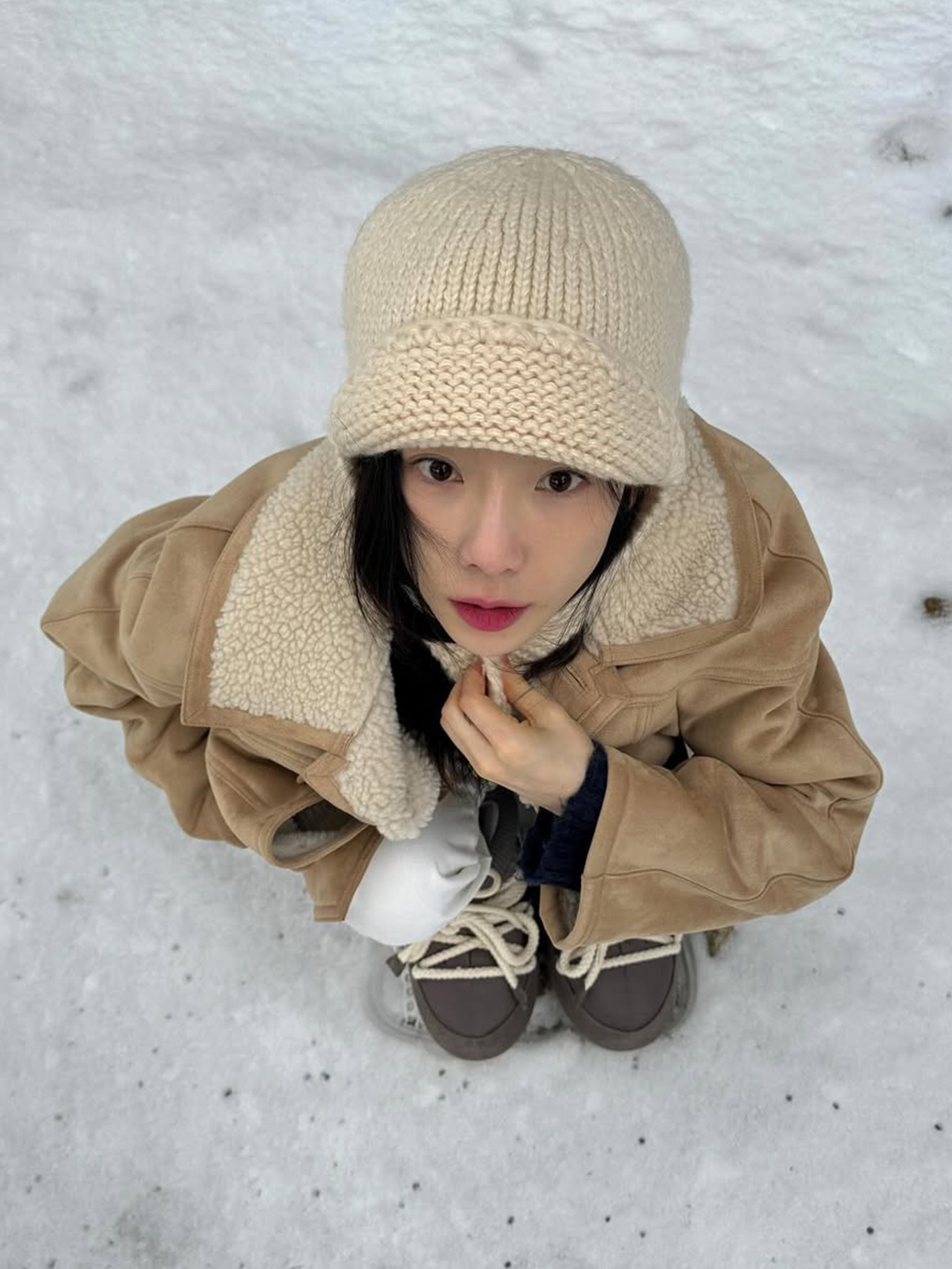 라이트 베이지색 니트 트루퍼햇을 쓰고 같은 톤의 스웨이드 무스탕을 입은 태연이 눈 밭에 앉아있는 사진